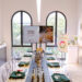 Bright fragrance shop workshop space with a long table set for a class, surrounded by product shelves and large arched windows outside light streaming in.